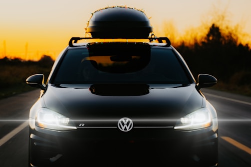 A black car with bright headlights is parked on a road during sunset. A luggage box is mounted on the roof, and decorative lights are wrapped around it. The sky is a gradient of orange, fading into darker hues towards the top.