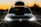 A black car with bright headlights is parked on a road during sunset. A luggage box is mounted on the roof, and decorative lights are wrapped around it. The sky is a gradient of orange, fading into darker hues towards the top.