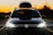 A black car with bright headlights is parked on a road during sunset. A luggage box is mounted on the roof, and decorative lights are wrapped around it. The sky is a gradient of orange, fading into darker hues towards the top.