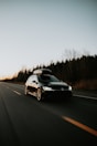 A black car with a roof box is traveling along a deserted highway, surrounded by bare trees and a clear sky at dusk.