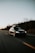 A black car with a roof box is traveling along a deserted highway, surrounded by bare trees and a clear sky at dusk.