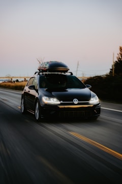 A black Volkswagen car with a roof box fixed on top, driving along an open road. The surrounding landscape includes blurred trees and fields, indicating motion. The road is wide with marked lanes, and the sky is clear, hinting at sunset or sunrise.
