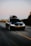 A black Volkswagen car with a roof box fixed on top, driving along an open road. The surrounding landscape includes blurred trees and fields, indicating motion. The road is wide with marked lanes, and the sky is clear, hinting at sunset or sunrise.