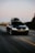A black Volkswagen car with a roof box fixed on top, driving along an open road. The surrounding landscape includes blurred trees and fields, indicating motion. The road is wide with marked lanes, and the sky is clear, hinting at sunset or sunrise.
