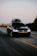 A black Volkswagen car with a roof box fixed on top, driving along an open road. The surrounding landscape includes blurred trees and fields, indicating motion. The road is wide with marked lanes, and the sky is clear, hinting at sunset or sunrise.