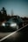 A dark-colored Volkswagen car is parked on a dimly lit road during dusk. The vehicle has its headlights on, and there's a cargo box on the roof decorated with small string lights. Trees and shrubs line the roadside under a clear, fading blue sky.