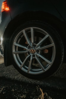 A close-up view of a car wheel with a prominent alloy rim, featuring a recognizable automotive logo at the center. The scene includes a dimly lit surrounding, with a hint of an orange taillight glowing in the background, indicating a nighttime or low-light setting.
