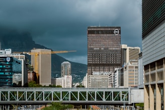 A cityscape featuring tall skyscrapers under a cloudy sky. In the foreground, a bridge crosses above a road. Construction cranes are visible among the buildings, suggesting ongoing development. Hills are visible in the background, partially obscured by low-hanging clouds.