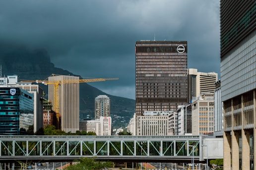 A cityscape featuring tall skyscrapers under a cloudy sky. In the foreground, a bridge crosses above a road. Construction cranes are visible among the buildings, suggesting ongoing development. Hills are visible in the background, partially obscured by low-hanging clouds.