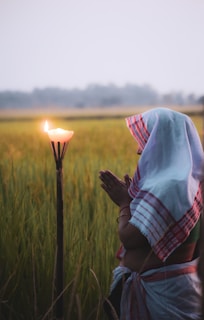 A person with their hands in a prayer position stands in a field, wearing traditional clothing. A lit candle on a holder is nearby, casting a warm glow. The landscape features tall grass and a soft-focused background with trees, enhanced by a serene evening light.