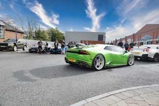 A gathering of people is observed around a collection of high-performance sports cars, including a vibrant green car prominently featured in the foreground. The scene takes place on a street lined with buildings and a clear, blue sky overhead.