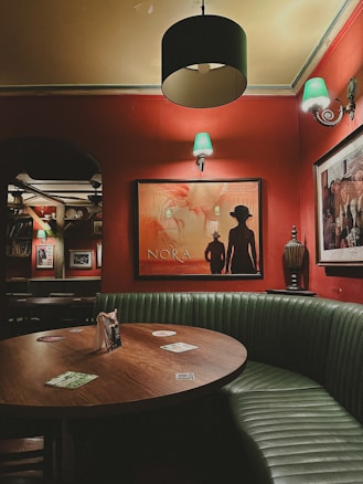A cozy, dimly lit interior of a bar or pub with red walls and various framed posters. A round wooden table is surrounded by green upholstered booth seating. Several colorful coasters are scattered on the table. The lighting is provided by wall sconces with green lampshades and a central black lamp, casting a warm ambiance.