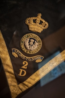 A dark fabric background displays an intricately embroidered gold crown, a circular emblem with the motto 'Nemo Me Impune Lacessit' around a symbol of a thistle, the phrase 'Second to None,' and the letters '2 D' in gold. The edges of the fabric are adorned with a detailed gold trim.