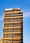 A tall, modern residential or commercial building with numerous floors, featuring a series of identical balconies on each floor. The building has a sign on top with the word 'parkmoza', framed against a clear blue sky. The sunlight casts a warm glow on the structure, highlighting its geometric design.