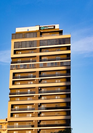 A tall, modern residential or commercial building with numerous floors, featuring a series of identical balconies on each floor. The building has a sign on top with the word 'parkmoza', framed against a clear blue sky. The sunlight casts a warm glow on the structure, highlighting its geometric design.