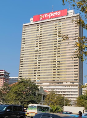 A tall building with a prominent red M-Pesa advertisement sign at the top. The structure has numerous windows and balconies. There are trees and several cars, including a white minivan, in the foreground. The sky is clear and blue.