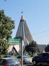 A unique, modern church with a tall, triangular structure and a cross at the peak, surrounded by lush greenery. Cars are parked nearby on a busy street labeled with a green sign that reads 'Avenida Armando Tivane.' Trees partially frame the scene, adding a touch of nature.