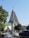 A unique, modern church with a tall, triangular structure and a cross at the peak, surrounded by lush greenery. Cars are parked nearby on a busy street labeled with a green sign that reads 'Avenida Armando Tivane.' Trees partially frame the scene, adding a touch of nature.