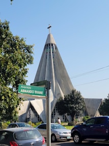 A unique, modern church with a tall, triangular structure and a cross at the peak, surrounded by lush greenery. Cars are parked nearby on a busy street labeled with a green sign that reads 'Avenida Armando Tivane.' Trees partially frame the scene, adding a touch of nature.