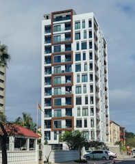 A modern high-rise building with a white facade and brown geometric accents. The building features several balconies with visible glass railings, and some have clothes hanging out. Surrounding the building are palm trees, a flag on a pole, and a small house with a red roof. A parked car and street elements are present in the foreground.