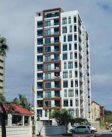 A modern high-rise building with a white facade and brown geometric accents. The building features several balconies with visible glass railings, and some have clothes hanging out. Surrounding the building are palm trees, a flag on a pole, and a small house with a red roof. A parked car and street elements are present in the foreground.