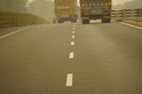 A two-lane road with a dashed white line in the center, flanked by trucks carrying cargo. The road extends into the distance with barriers on either side, and the atmosphere appears hazy, possibly due to fog or dust.