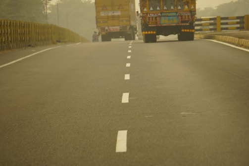 A two-lane road with a dashed white line in the center, flanked by trucks carrying cargo. The road extends into the distance with barriers on either side, and the atmosphere appears hazy, possibly due to fog or dust.