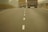 A two-lane road with a dashed white line in the center, flanked by trucks carrying cargo. The road extends into the distance with barriers on either side, and the atmosphere appears hazy, possibly due to fog or dust.