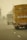 Several trucks are seen driving along a foggy highway. The back of a prominent yellow truck is visible with some signage and markings. The road has marked lanes, and the fog creates a hazy atmosphere, obscuring the distance.