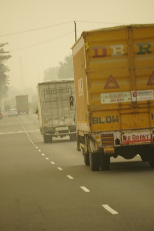 Several trucks are seen driving along a foggy highway. The back of a prominent yellow truck is visible with some signage and markings. The road has marked lanes, and the fog creates a hazy atmosphere, obscuring the distance.