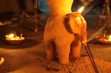 A clay sculpture resembling an abstract figure is surrounded by burning candles and incense sticks placed in small bowls. The warm glow creates a serene and spiritual atmosphere, with shadows and smoke gently wafting through the scene. There is also a pile of grains or seeds near the base of the sculpture.