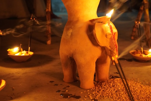 A clay sculpture resembling an abstract figure is surrounded by burning candles and incense sticks placed in small bowls. The warm glow creates a serene and spiritual atmosphere, with shadows and smoke gently wafting through the scene. There is also a pile of grains or seeds near the base of the sculpture.