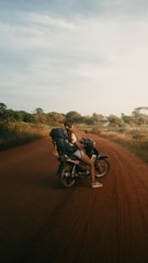 A person with a backpack sits on a motorcycle on a dirt road surrounded by grass and trees under a vast, slightly cloudy sky. The scene conveys a sense of exploration and travel.