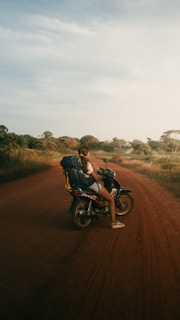 A person with a backpack sits on a motorcycle on a dirt road surrounded by grass and trees under a vast, slightly cloudy sky. The scene conveys a sense of exploration and travel.