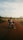 A person with a backpack sits on a motorcycle on a dirt road surrounded by grass and trees under a vast, slightly cloudy sky. The scene conveys a sense of exploration and travel.
