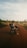 A person with a backpack sits on a motorcycle on a dirt road surrounded by grass and trees under a vast, slightly cloudy sky. The scene conveys a sense of exploration and travel.
