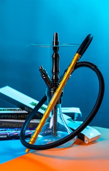 A hookah with a golden and black hose is prominently placed against a vibrant blue background. Books with titles related to 'flipping houses' and colorful packaging are casually arranged around the base.