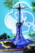 A blue hookah with geometric patterns sits in the center, surrounded by green leaves. It is placed on a colorful mat with 'CHIMBEROS' text. Black charcoal cubes are scattered around, and a mustache-shaped accessory is also present. The background features neon blue and white light in a circular design.