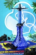 A blue hookah with geometric patterns sits in the center, surrounded by green leaves. It is placed on a colorful mat with 'CHIMBEROS' text. Black charcoal cubes are scattered around, and a mustache-shaped accessory is also present. The background features neon blue and white light in a circular design.