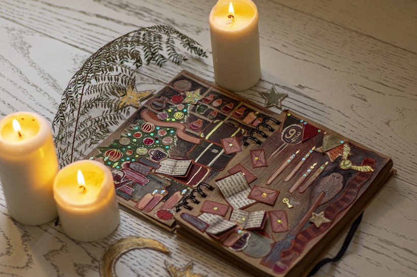 A wooden table is beautifully decorated with three lit white candles, adding a warm glow. An open scrapbook displays various colorful and thematic elements including envelopes, potion bottles, and magical motifs. Glittery stars and decorative ferns accentuate the setup, suggesting a magical or whimsical theme.