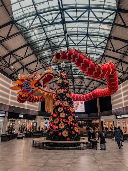 A large shopping mall decorated for a festive occasion with a giant dragon sculpture hanging above a Christmas tree. The tree is adorned with red and gold ornaments and Chinese zodiac symbols. The mall has a high, glass-panelled ceiling, and various stores, including a Samsung and a Karl Lagerfeld shop, can be seen in the background. Shoppers, including a family with children, are walking around on the floor.