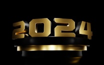 Large, bold metallic numbers in gold color with a shiny finish are placed prominently against a dark background, forming the year 2024. The numbers appear three-dimensional and are set above a dark pedestal, adding a dramatic effect.