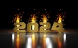 Golden numbers '2024' are displayed prominently against a dark background. Behind the numbers, bright and vibrant fireworks burst upwards, casting a warm glow and reflection on the surface below.