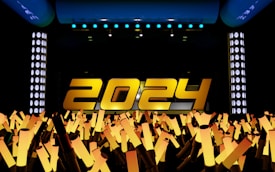Large yellow numbers spelling out '2024' are prominently displayed on a stage with bright lights. Below the numbers, a crowd of illuminated light sticks points upward, creating a celebratory atmosphere.