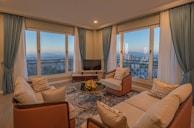 A cozy living room features elegant blue curtains framing large windows that offer a panoramic city view. Earth-toned sofas and armchairs are arranged around a glass coffee table with a floral centerpiece. Warm lighting creates a serene ambiance, complemented by a plush area rug.
