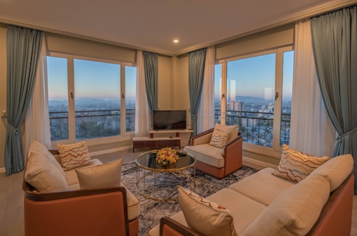 A cozy living room features elegant blue curtains framing large windows that offer a panoramic city view. Earth-toned sofas and armchairs are arranged around a glass coffee table with a floral centerpiece. Warm lighting creates a serene ambiance, complemented by a plush area rug.