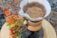 A pour-over coffee maker holds fresh coffee with bubbles forming on the surface. It rests on a wooden surface surrounded by autumn leaves and red berries. The scene suggests an outdoor setting, possibly in a forest or garden.