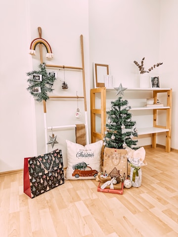 A cozy, festive indoor scene featuring a small decorated Christmas tree with silver ornaments. Nearby, there is a pillow with a car and Christmas tree graphic, a holiday gift bag, a decorative ladder with a hanging wreath and rainbow, several wrapped gifts, and a wooden shelf with framed pictures and a vase.