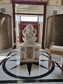 A large white statue of a seated elephant-headed deity, likely Ganesha. The statue is intricately detailed and positioned on a decorative pedestal with ornate carvings. Incense burns to the side, creating a serene atmosphere. The floor is adorned with a geometric pattern, and a donation box is visible nearby.