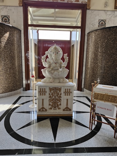 A large white statue of a seated elephant-headed deity, likely Ganesha. The statue is intricately detailed and positioned on a decorative pedestal with ornate carvings. Incense burns to the side, creating a serene atmosphere. The floor is adorned with a geometric pattern, and a donation box is visible nearby.